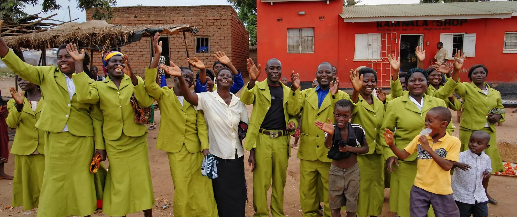 A group of people (women, men and children) of an African cooperative is waving their hands to the camera, standing in front of a red brick house.