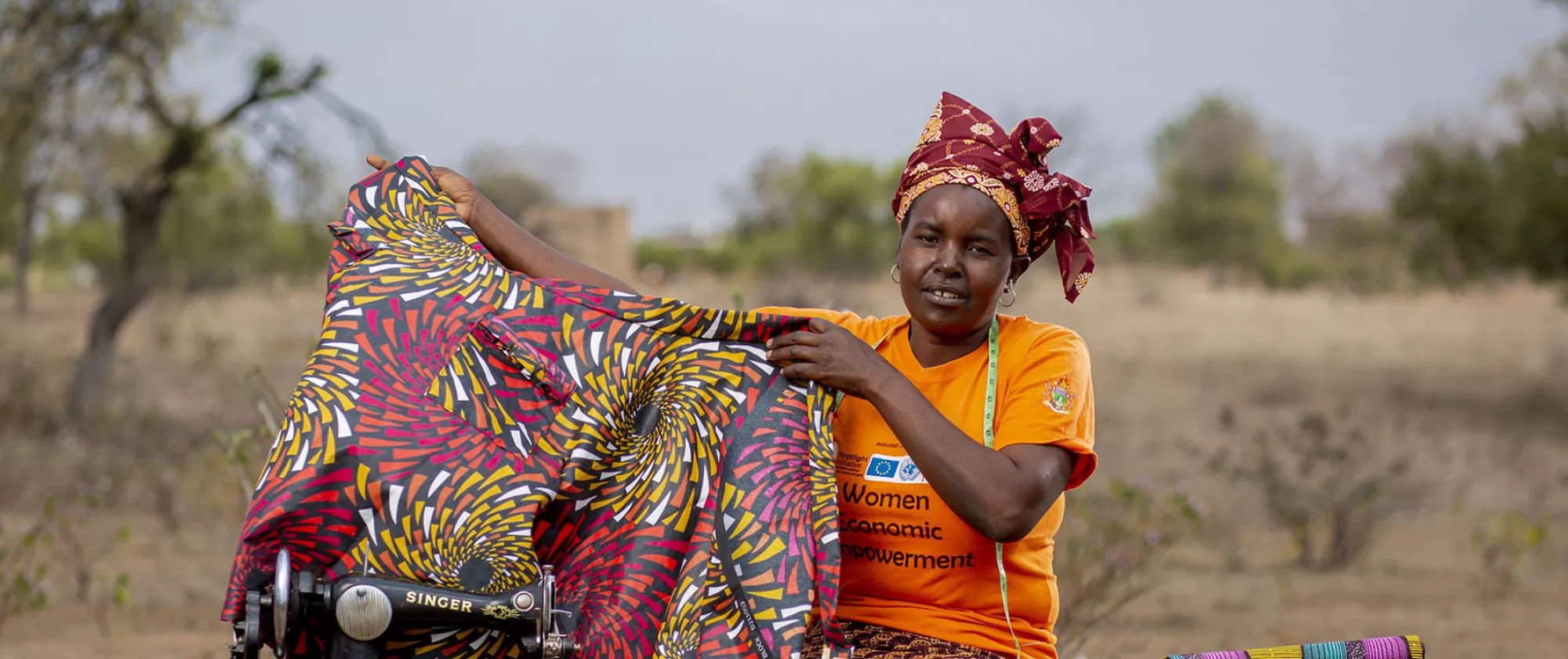 Zimbabwean fashion designer Martha Simango sits outside by a field and holds up one of her creations, a multicoloured dress she is working on