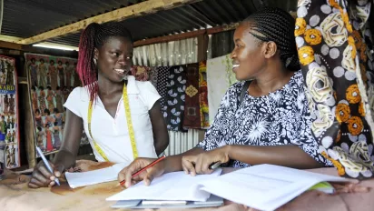 Two African women entrepreneurs are discussing a document. A cloth stand is in the background.